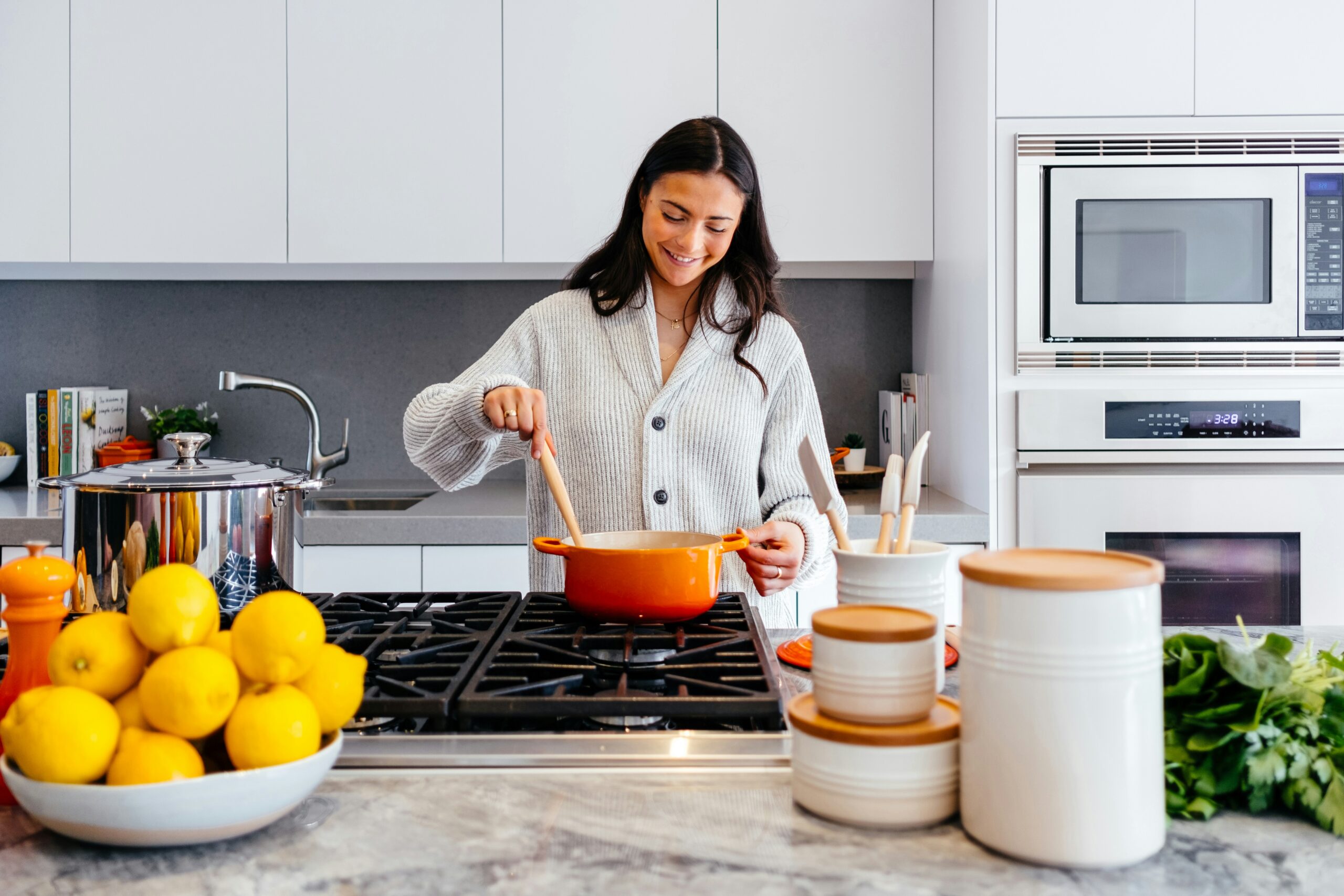 A woman cooking at a gas stove with an orange pot in a modern kitchen, surrounded by lemons, containers, and greens.