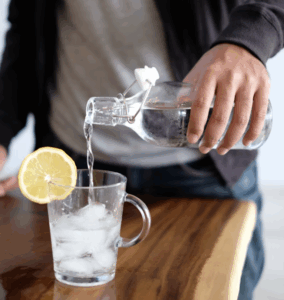 Person pouring water into a glass mug with ice and a lemon slice.