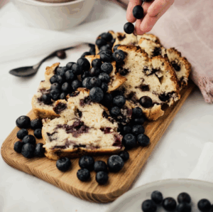 Slices of blueberry bread on a wooden board with scattered blueberries and a hand adding more.