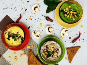 Three bowls of colorful dips on red, yellow, and green plates, garnished with herbs and seeds, surrounded by mushrooms, chili peppers, and bread slices.