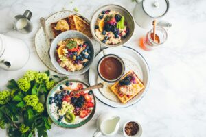 Breakfast spread with oatmeal, waffles, yogurt, a latte, a teapot, and fresh flowers on a marble surface.