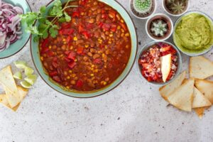 A bowl of chili surrounded by accompaniments including sliced onions, salsa, guacamole, and lime wedges, alongside small pots with cacti.
