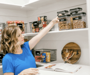 A woman in a blue shirt reaches for a labeled container in a neatly organized pantry with white shelves, wire baskets, and an open cookbook.