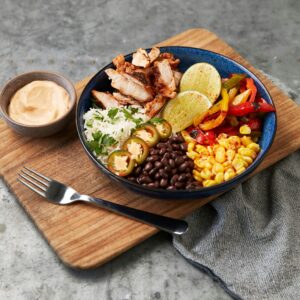 A vibrant bowl of chicken, rice, beans, peppers, corn, lime, and jalapeños on a cutting board with a side of dipping sauce and a fork.