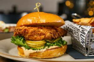 Close-up of a fried chicken sandwich with lettuce, pickles, and sauce on a brioche bun, next to a basket of chips.
