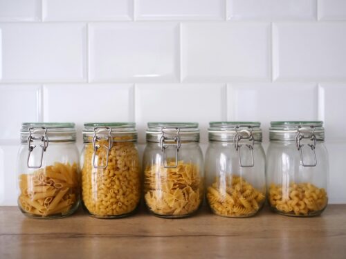 Five glass jars filled with different types of pasta against a white tiled backsplash.