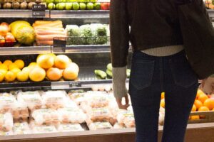 Person standing in front of a supermarket produce display with fruits, vegetables, and eggs.