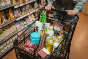 A grocery cart with carrots, proteins, apples, salami, milk, Zevia soda cans, soap, and other items in a store aisle.
