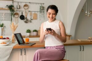 A woman in a white top and pink-striped pants sits on a kitchen counter, smiling at her smartphone. The countertop has fruit, orange juice, and a laptop.