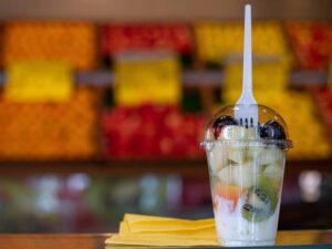 A plastic cup filled with mixed fruits and yogurt with a fork stuck through the lid, against a blurred background of colorful produce.