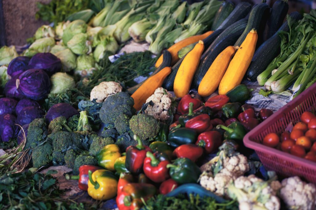 Assorted fresh vegetables displayed at a market, including cabbage, broccoli, cauliflower, bell peppers, zucchini, squash, celery, and cherry tomatoes.