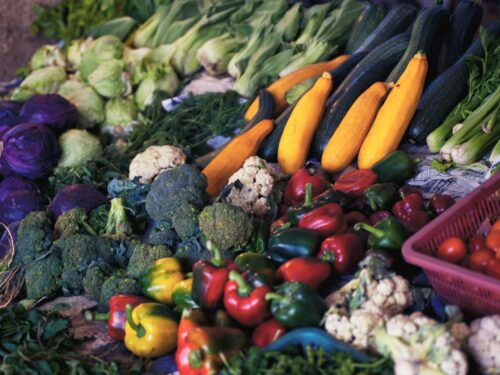 Assorted fresh vegetables displayed at a market, including cabbage, broccoli, cauliflower, bell peppers, zucchini, squash, celery, and cherry tomatoes.