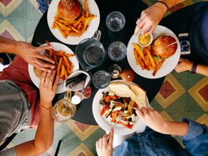 Overhead view of a table with hamburgers, fries, and grilled vegetables on plates, with hands reaching for food.