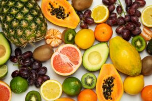 A colorful assortment of various fruits including pineapple, papaya, grapefruit, citrus fruits, grapes, avocado, and kiwi on a white background.