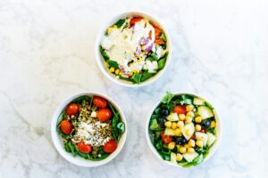 Three bowls of salad with various ingredients like spinach, tomatoes, chickpeas, and feta, arranged on a marble surface.