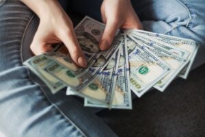 Hands holding a spread of $100 U.S. dollar bills over denim-clad knees.