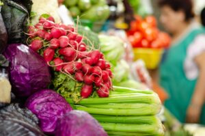 A colorful market display with radishes, celery, and purple cabbage in focus. Blurred background with tomatoes and a person.