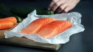 Two raw salmon fillets on parchment paper with carrots and a zucchini nearby.