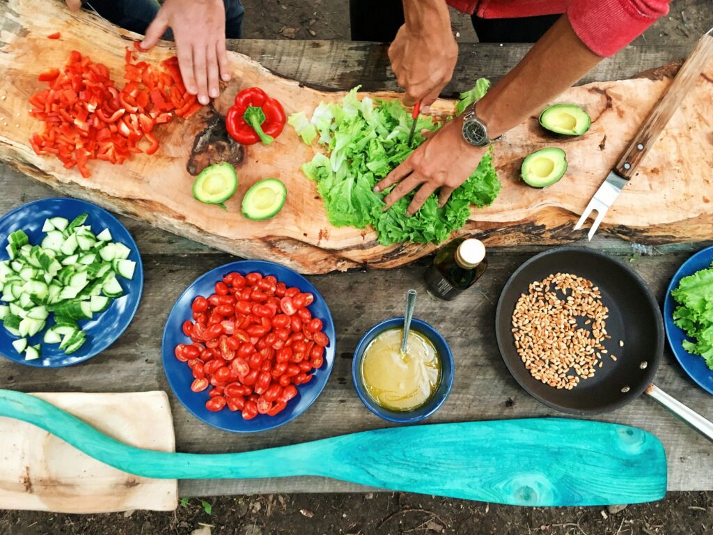 Top view of a wooden table with fresh vegetables and kitchen items being prepared while two people cut them together.