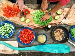 Top view of a wooden table with fresh vegetables and kitchen items being prepared while two people cut them together.
