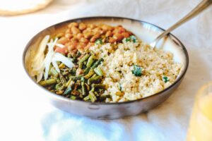 A bowl with quinoa, pinto beans, seasoned greens, and shaved strips of cheese, with a spoon on the side.