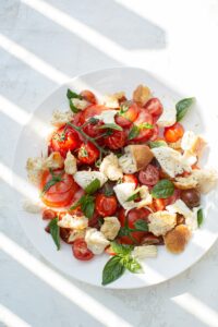 A colorful salad with tomatoes, basil leaves, mozzarella cheese, and bread pieces on a white plate, casting diagonal shadows on a textured surface.