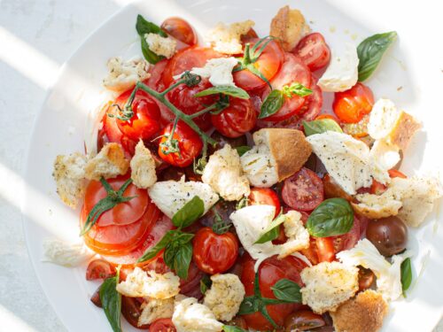 A colorful salad with tomatoes, basil leaves, mozzarella cheese, and bread pieces on a white plate, casting diagonal shadows on a textured surface.