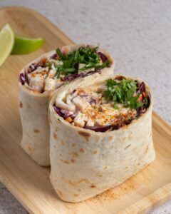 Two burritos filled with shredded cabbage, chicken, and cilantro on a wooden cutting board, accompanied by a wedge of lime.