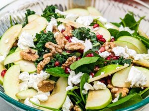 A colorful salad with kale, spinach, green apple slices, pomegranate seeds, walnuts, and crumbled cheese in a clear glass bowl.